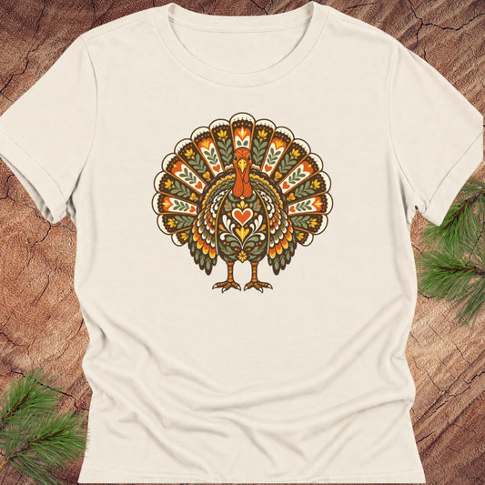 An off-white short-sleeve crewneck t-shirt featuring a colorful, vintage-style graphic of a turkey with a heart on its chest, laid flat on a rustic wood background with pine needles.