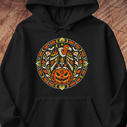 A black pullover hoodie with a large circular Halloween graphic on the chest, featuring a carved pumpkin, eagle, and autumn floral design in orange, yellow, white, and black, shown against a wood-textured background.
