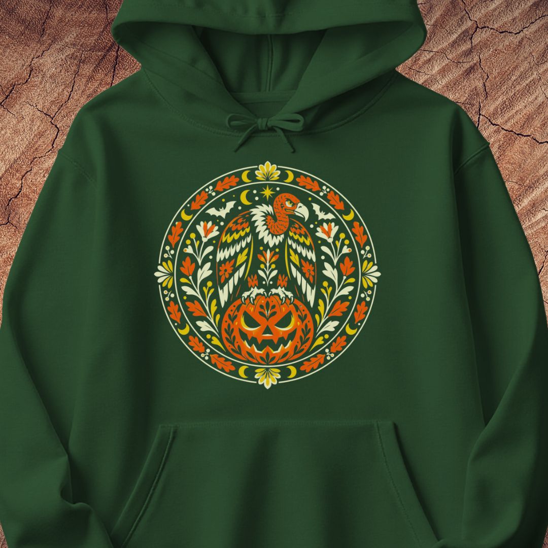 A dark green pullover hoodie featuring a large circular graphic on the chest with a stylized bird, pumpkin, and floral motifs in orange, yellow, white, and green, displayed against a textured brown wood background.