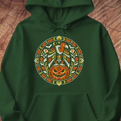 A dark green pullover hoodie featuring a large circular graphic on the chest with a stylized bird, pumpkin, and floral motifs in orange, yellow, white, and green, displayed against a textured brown wood background.