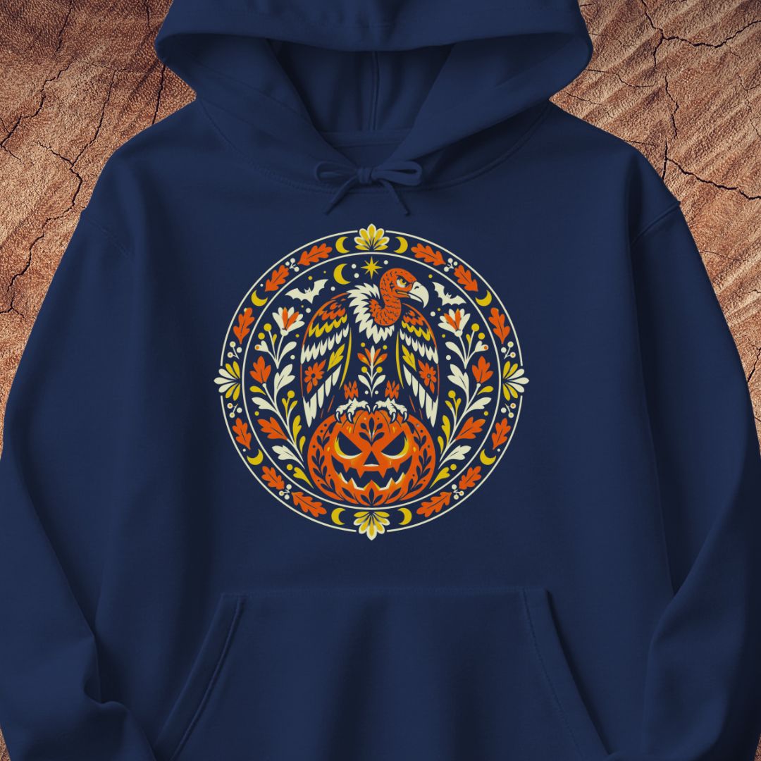 A navy blue pullover hoodie featuring a large, colorful circular graphic on the chest with a stylized eagle, pumpkin, and floral motifs in orange, yellow, white, and green, displayed against a textured brown wood background.