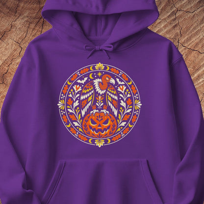 A purple pullover hoodie featuring a large circular graphic on the chest with a stylized eagle and carved pumpkin in orange, yellow, and white, shown against a wood-textured background.