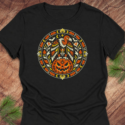 A black short-sleeve crewneck t-shirt featuring a circular graphic print of a bald eagle perched on a carved jack-o'-lantern pumpkin, surrounded by autumn leaves and stars. The shirt is laid flat on a rustic wood background with pine needles in the corners.