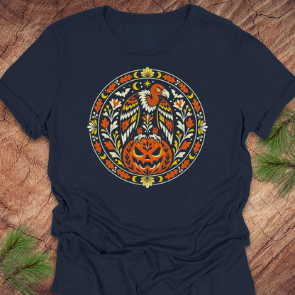 A navy blue short-sleeve t-shirt with a circular graphic of a bald eagle perched on a carved jack-o'-lantern pumpkin, surrounded by autumn leaves and a crescent moon, laid flat on a rustic wood background with pine branches.