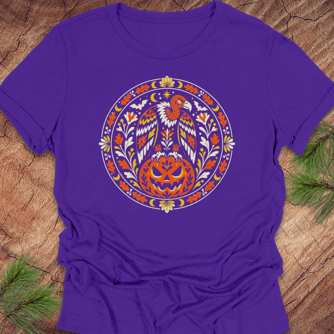 A purple short-sleeve crewneck t-shirt featuring a colorful circular graphic of a bald eagle and a jack-o'-lantern pumpkin, laid flat on a rustic wood background with pine needles in the corners.