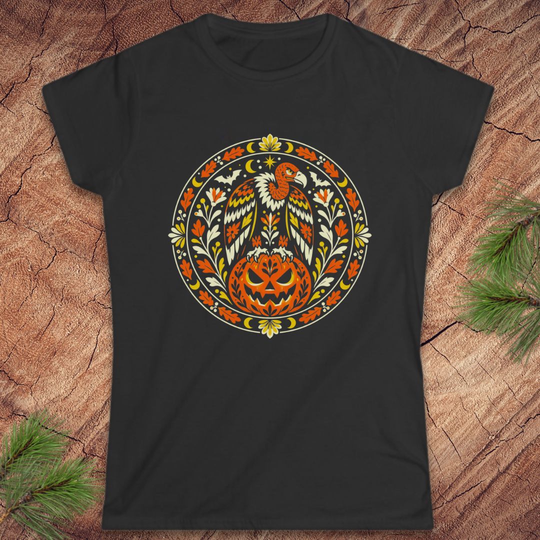 A black short-sleeve crewneck t-shirt featuring a circular folk-art graphic of a bald eagle and a carved jack-o'-lantern pumpkin, displayed flat on a rustic wood background with pine branches.