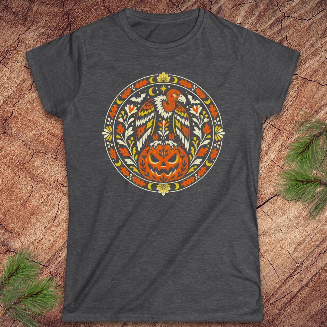 A dark heather gray t-shirt featuring a circular folk-art style graphic of a bald eagle perched on a carved jack-o'-lantern pumpkin, surrounded by floral and leaf motifs. The t-shirt is displayed flat on a textured wood background with pine branches in the corners.