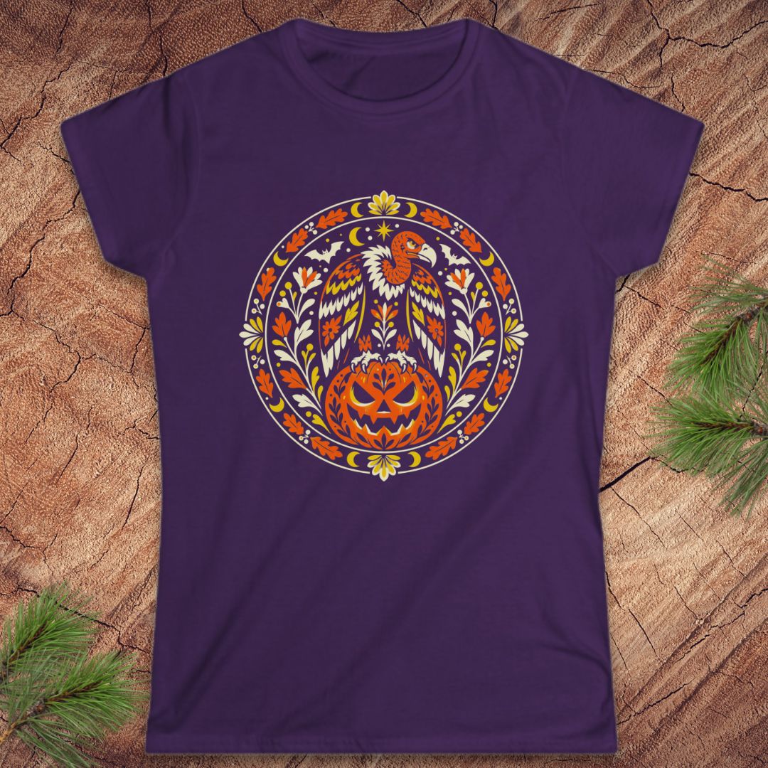 A purple short-sleeve crewneck t-shirt featuring a circular graphic of a bald eagle perched on a carved jack-o'-lantern pumpkin, surrounded by autumn leaves. The shirt is displayed flat on a textured wood background with pine branches in the corners.