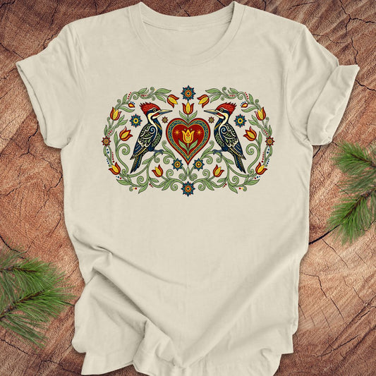 Beige t-shirt with a design of two woodpeckers around a heart on a wooden surface.