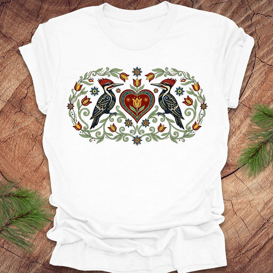 White t-shirt with a decorative design of two birds and a heart on a wooden surface.