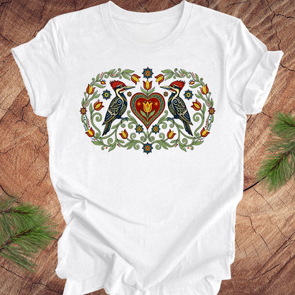White t-shirt with a design of two woodpeckers around a heart on a wooden surface.
