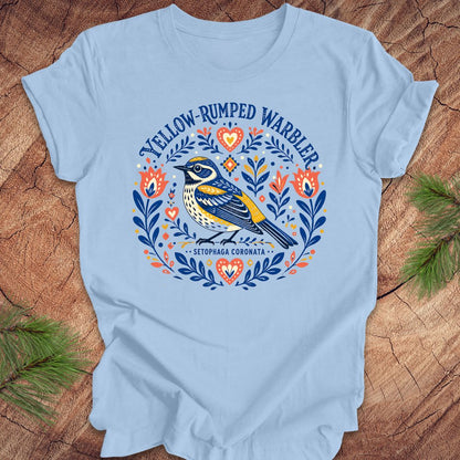 Blue t-shirt with a folk art yellow rumped warbler design on a wood background.