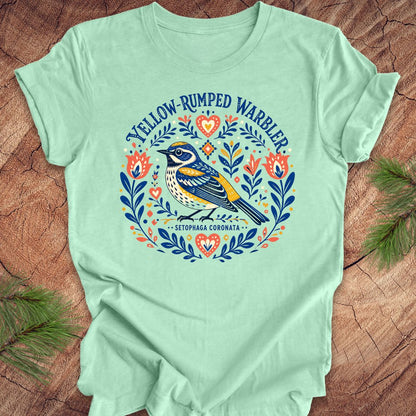Mint green t-shirt with a folk art yellow rumped warbler design on a wood background.