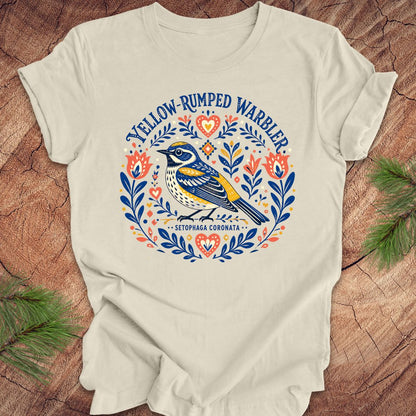 Beige t-shirt with a folk art yellow rumped warbler design on a wood background.