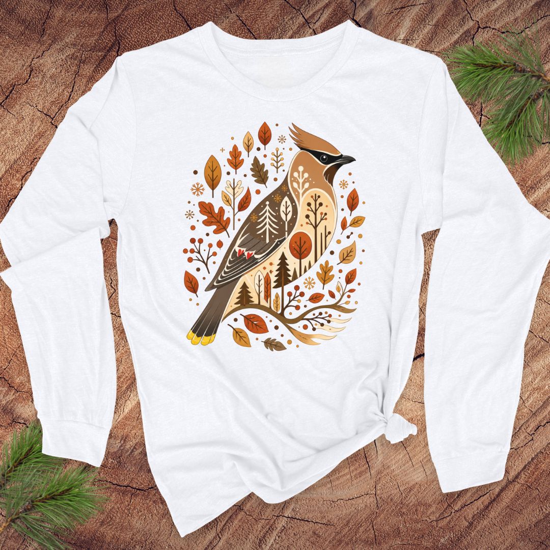 A white long-sleeve t-shirt with a graphic of a cedar waxbird surrounded by autumn leaves and trees, laid flat on a rustic wood background with pine branches.