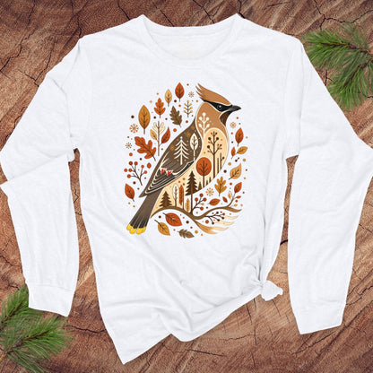 A white long-sleeve t-shirt with a graphic of a cedar waxbird surrounded by autumn leaves and trees, laid flat on a rustic wood background with pine branches.