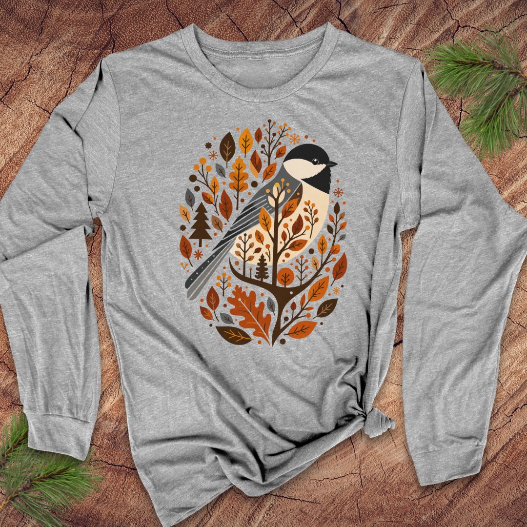 A heather gray long-sleeve t-shirt with a graphic of a black-capped chickadee surrounded by autumn leaves and pine trees, laid flat on a rustic wood background with pine needles.