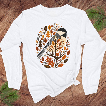 A white long-sleeve t-shirt with a graphic of a chickadee surrounded by autumn leaves, styled flat on a rustic wood background with pine branches.