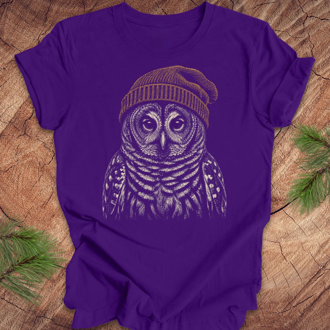 Purple t-shirt with a barred owl wearing a beanie on a wooden surface.