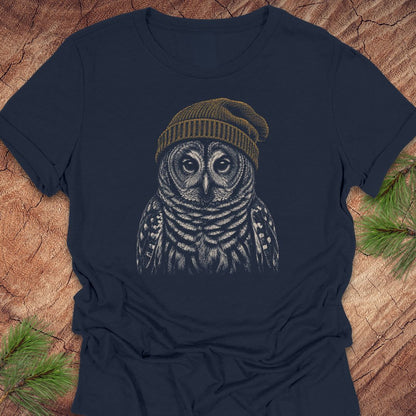 A navy blue short-sleeve t-shirt with a detailed graphic of an owl wearing a brown knit beanie, laid flat on a rustic wood background with pine needles in the corners.