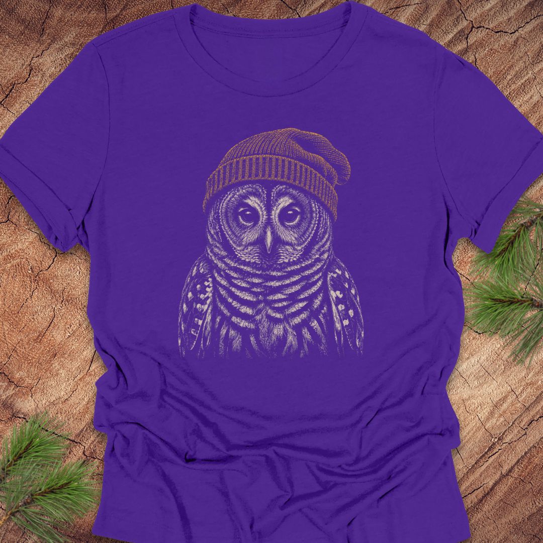 A purple short-sleeve crewneck t-shirt featuring a graphic of an owl wearing a knit beanie, laid flat on a rustic wood background with pine needles in the corners.