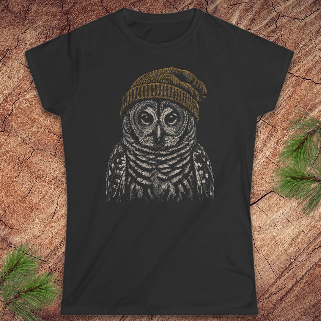 A black short-sleeve crewneck t-shirt featuring a detailed graphic of a barred owl wearing a brown knit beanie, displayed flat on a rustic wood background with pine needles.