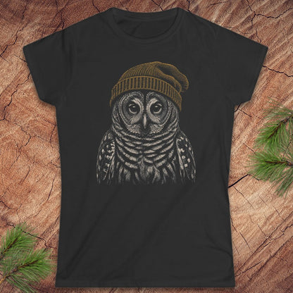 A black short-sleeve crewneck t-shirt featuring a detailed graphic of a barred owl wearing a brown knit beanie, displayed flat on a rustic wood background with pine needles.