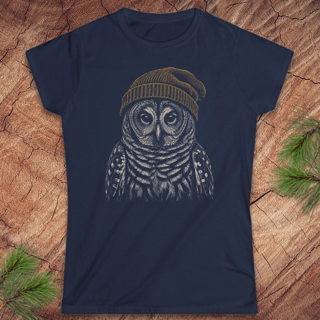 A navy blue short-sleeve crewneck t-shirt featuring a detailed graphic of an owl wearing a brown knit beanie, laid flat on a textured wood background with pine branches in the corners.