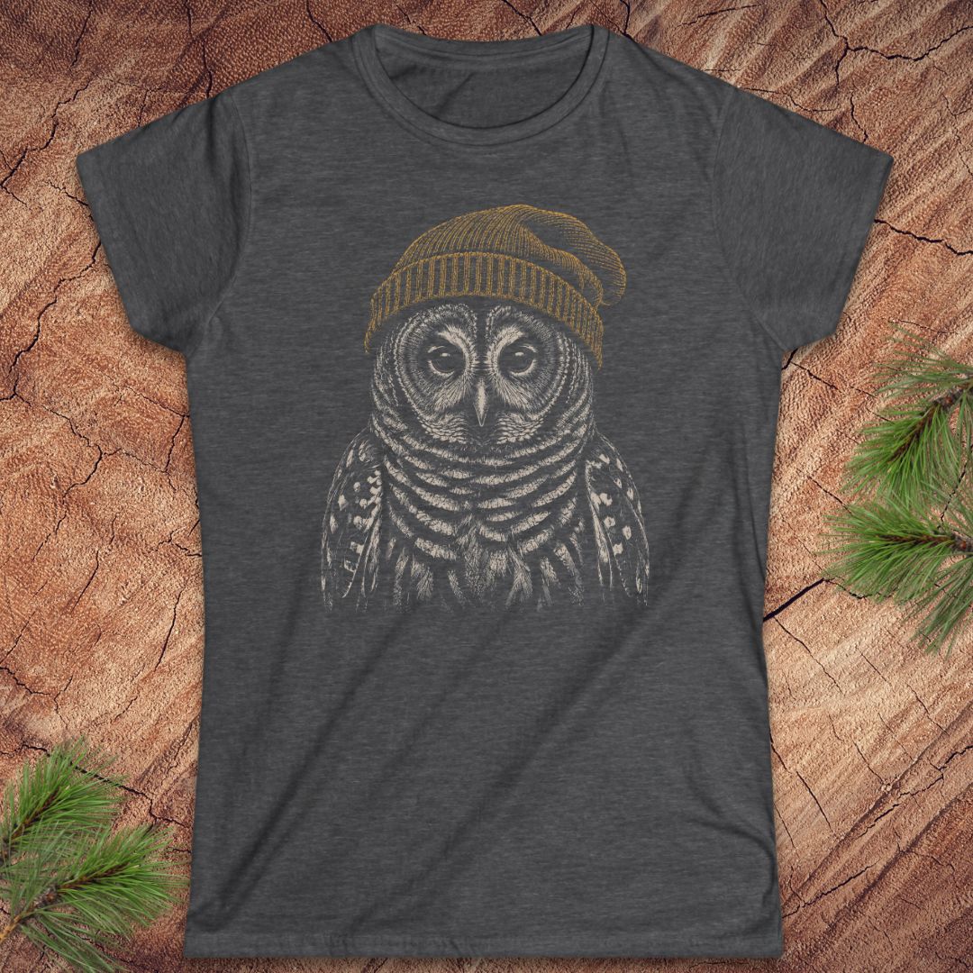 A dark heather gray t-shirt featuring a graphic of an owl wearing a yellow beanie, laid flat on a rustic wood background with pine branches in the corners.