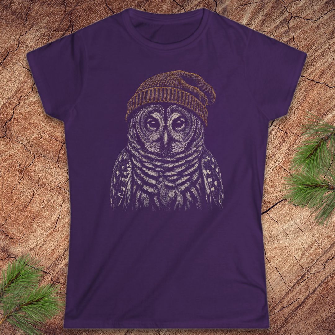A purple short-sleeve crewneck t-shirt featuring a detailed graphic of a owl wearing a knit beanie, laid flat on a rustic wood background with pine needles in the corners.
