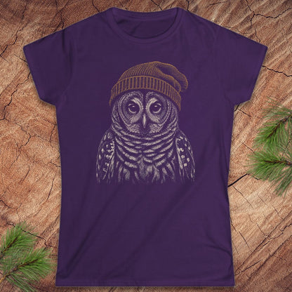 A purple short-sleeve crewneck t-shirt featuring a detailed graphic of a owl wearing a knit beanie, laid flat on a rustic wood background with pine needles in the corners.