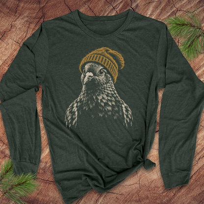 A dark forest green long-sleeve t-shirt featuring a graphic print of a snowy owl wearing a mustard yellow knit beanie, laid flat on a textured wood background with pine needles.