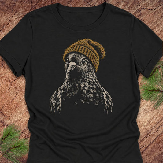 A black short-sleeve t-shirt featuring a graphic print of a pigeon wearing a mustard yellow knit beanie, laid flat on a rustic wood background with pine needles in the corners.