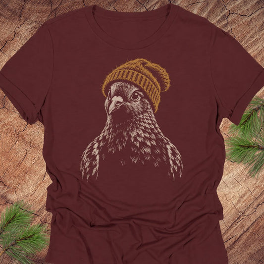 A dark maroon short-sleeve t-shirt featuring a graphic of a pigeon wearing a yellow knit beanie, laid flat on a rustic wood background with pine needles in the corners.