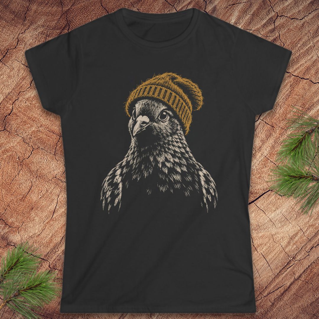 A black short-sleeve t-shirt featuring a graphic print of a falcon wearing a mustard yellow knit beanie, laid flat on a rustic wood background with pine needles in the corners.