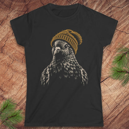 A black short-sleeve t-shirt featuring a graphic print of a falcon wearing a mustard yellow knit beanie, laid flat on a rustic wood background with pine needles in the corners.