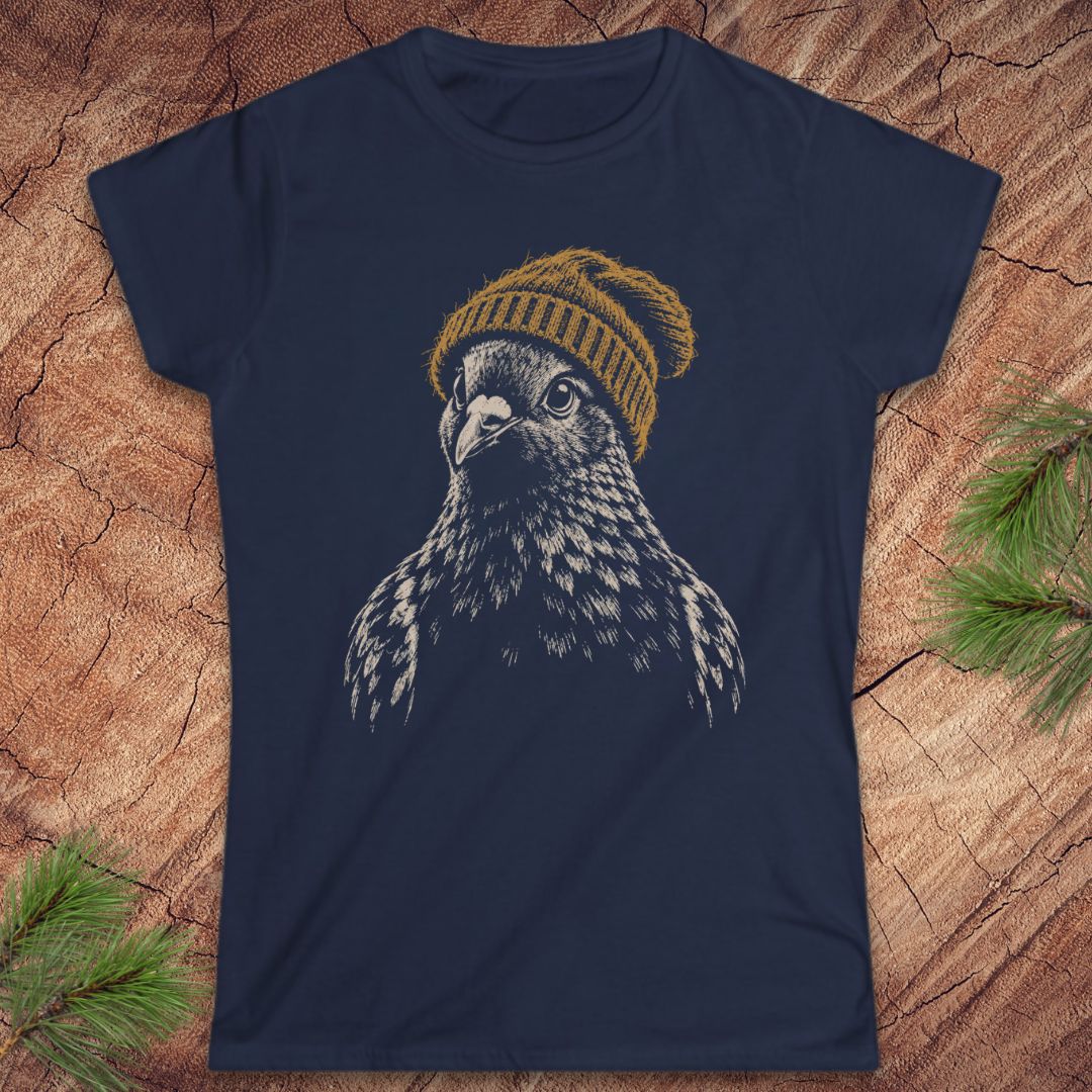 A navy blue short-sleeve t-shirt featuring a graphic of a bird of prey wearing a mustard yellow knit beanie, laid flat on a rustic wood background with pine branches in the corners.