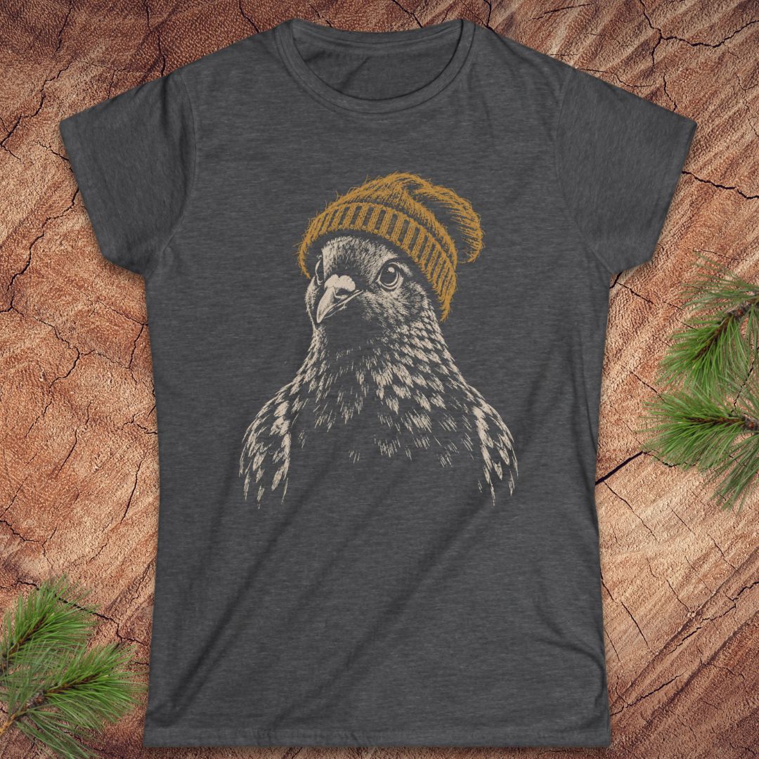 A dark heather gray t-shirt featuring a graphic print of a falcon wearing a yellow knit beanie, laid flat on a rustic wood background with pine needles in the corners.