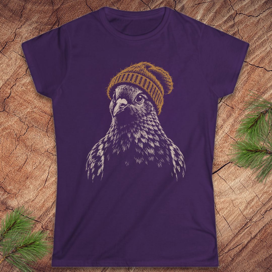 A purple short-sleeve crewneck t-shirt featuring a graphic of a bird of prey wearing a yellow knit beanie, laid flat on a textured wood background with pine branches in the corners.