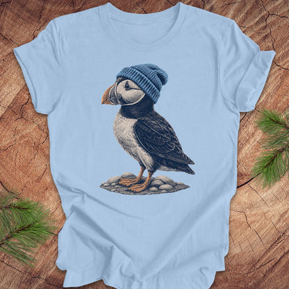 Light blue t-shirt with a puffin wearing a blue beanie on a wooden surface.