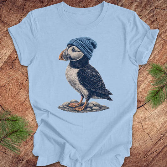 Light blue t-shirt with a puffin wearing a blue beanie on a wooden surface.