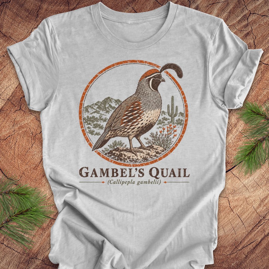 A light gray short-sleeve t-shirt with a 'Gambel's Quail' graphic print, laid flat on a rustic wood background with pine branches in the corners.