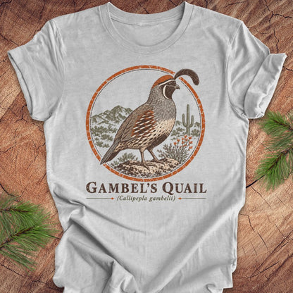A light gray short-sleeve t-shirt with a 'Gambel's Quail' graphic print, laid flat on a rustic wood background with pine branches in the corners.