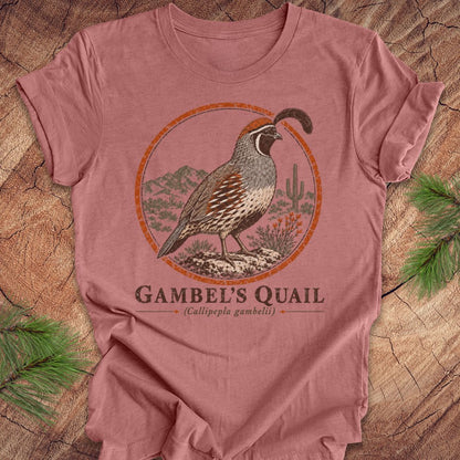 A dusty rose short-sleeve t-shirt featuring a vintage-style graphic of a Gambel's Quail in a desert landscape, laid flat on a rustic wood background with pine branches.