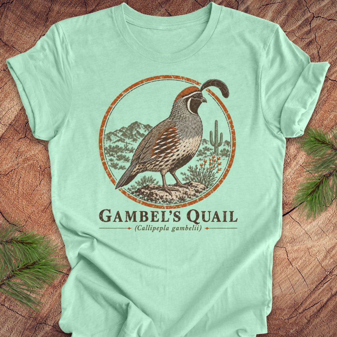 A mint green short-sleeve t-shirt with a detailed graphic of a Gambel's Quail bird inside a circular desert landscape design, laid flat on a rustic wood background with pine needles.