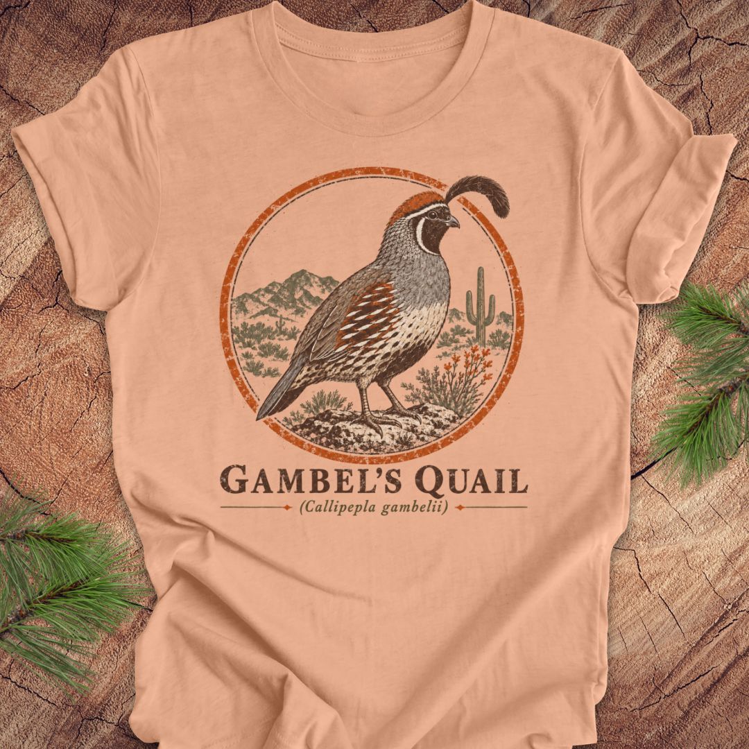 A peach-colored short-sleeve t-shirt with a vintage-style illustration of a Gambel's Quail in a desert landscape, displayed flat on a rustic wood background with pine needles.