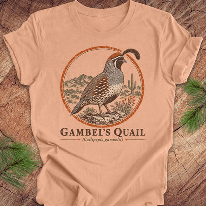 A peach-colored short-sleeve t-shirt with a vintage-style illustration of a Gambel's Quail in a desert landscape, displayed flat on a rustic wood background with pine needles.
