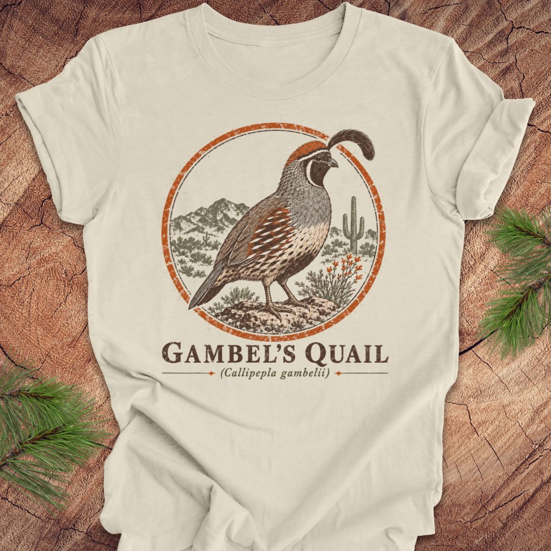 An off-white short-sleeve t-shirt with a vintage-style Gambel's Quail graphic, laid flat on a rustic wood background with pine needles in the corners.