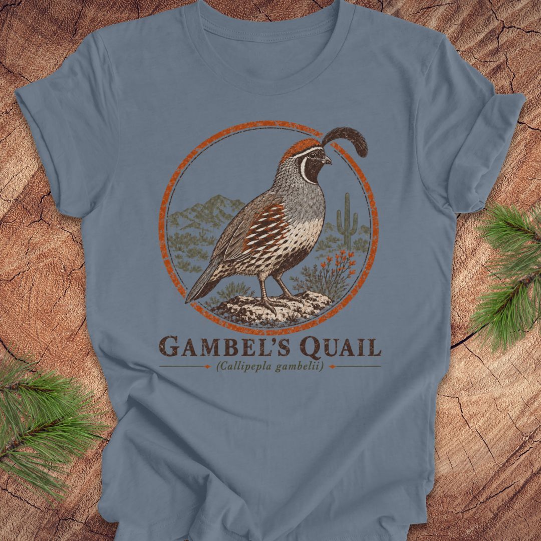 A slate blue crewneck t-shirt featuring a vintage-style graphic of a Gambel's Quail bird inside an orange circular frame, with 'GAMBEL'S QUAIL' printed below. The shirt is displayed flat on a rustic wood background with pine needles in the corners.