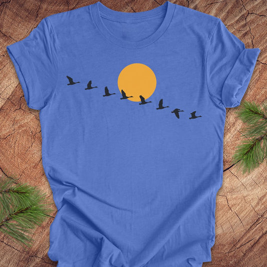 A blue short-sleeve crewneck t-shirt laid flat on a rustic wood background with pine branches in the corners. The shirt features a graphic of a large yellow-orange sun with a line of black flying birds in front of it.
